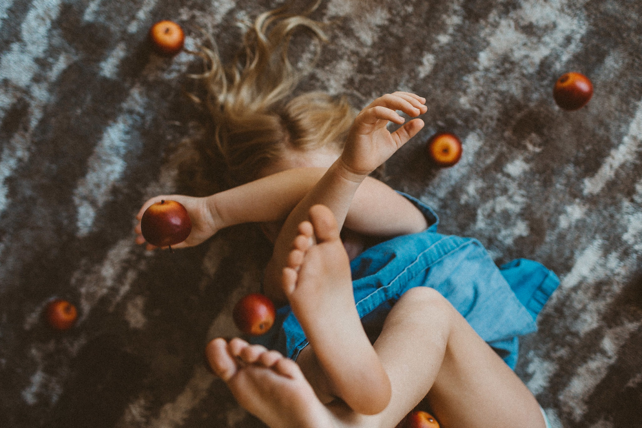 girl laying on carpeted floor, surrounded by apples and covering her face with her arms