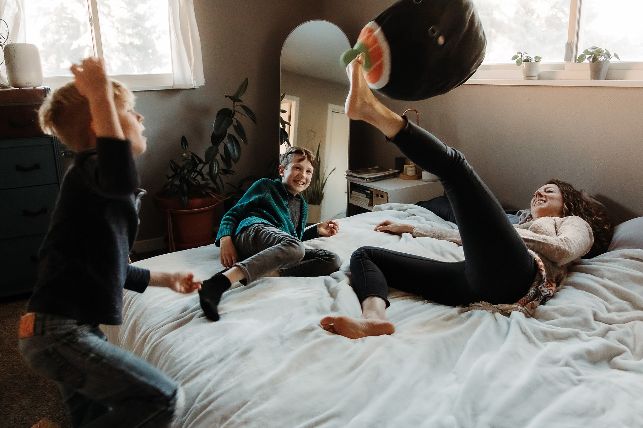 mother and kids play on bed in room of their home as they kick pillows around, smiling and laughing