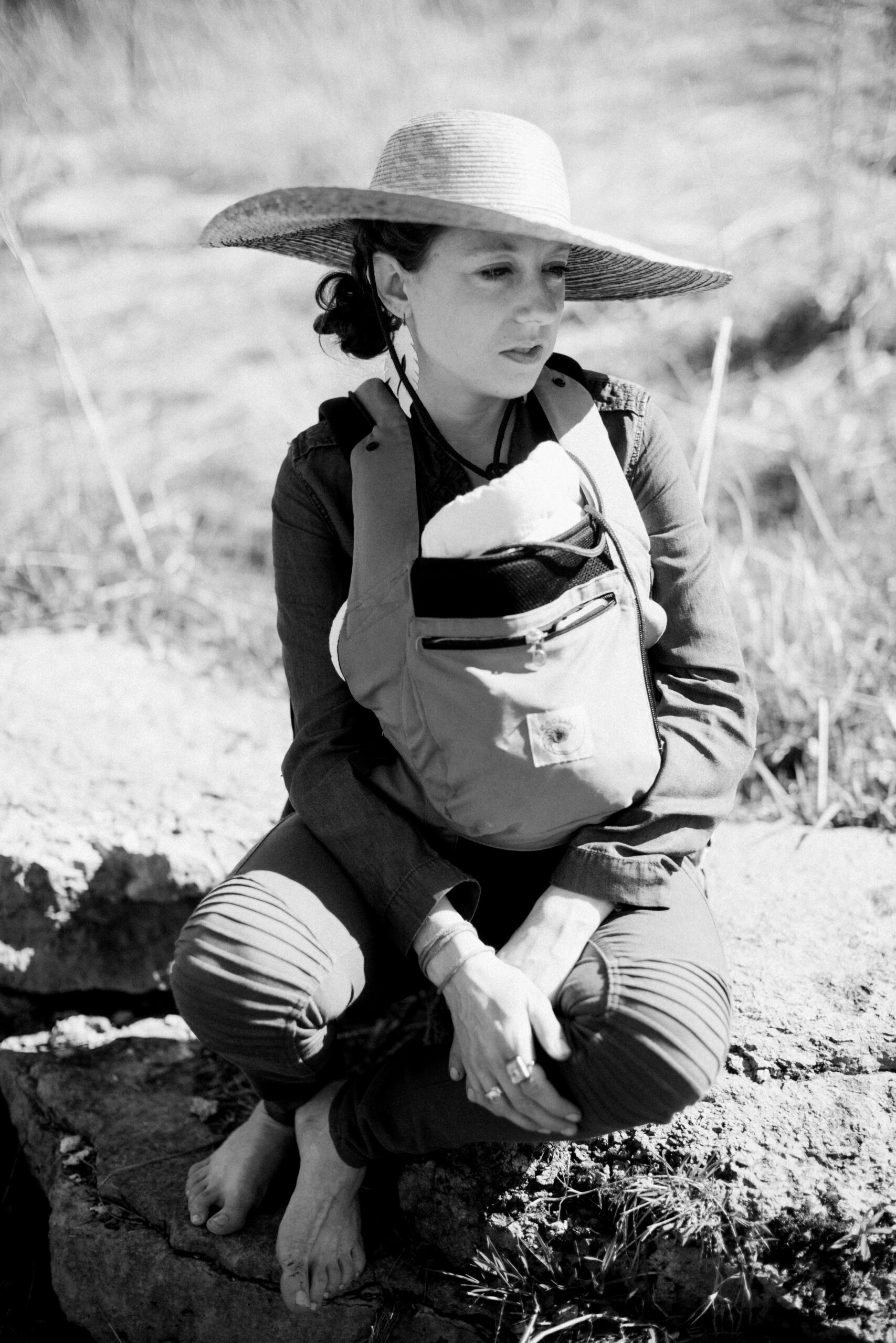 woman sitting in field by creek in colorado with wide sunhat on, carrying baby in a carrier