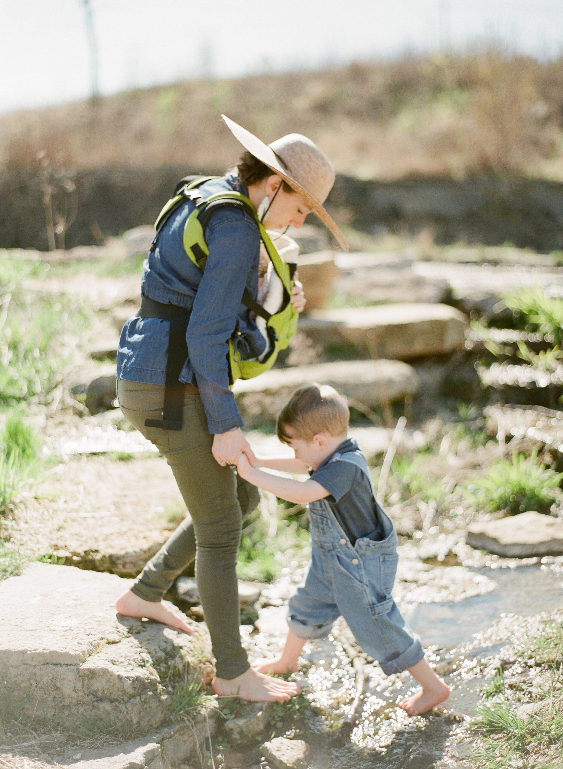 Barefoot mother helps barefoot child step across creek in Boulder while holding baby in carrier