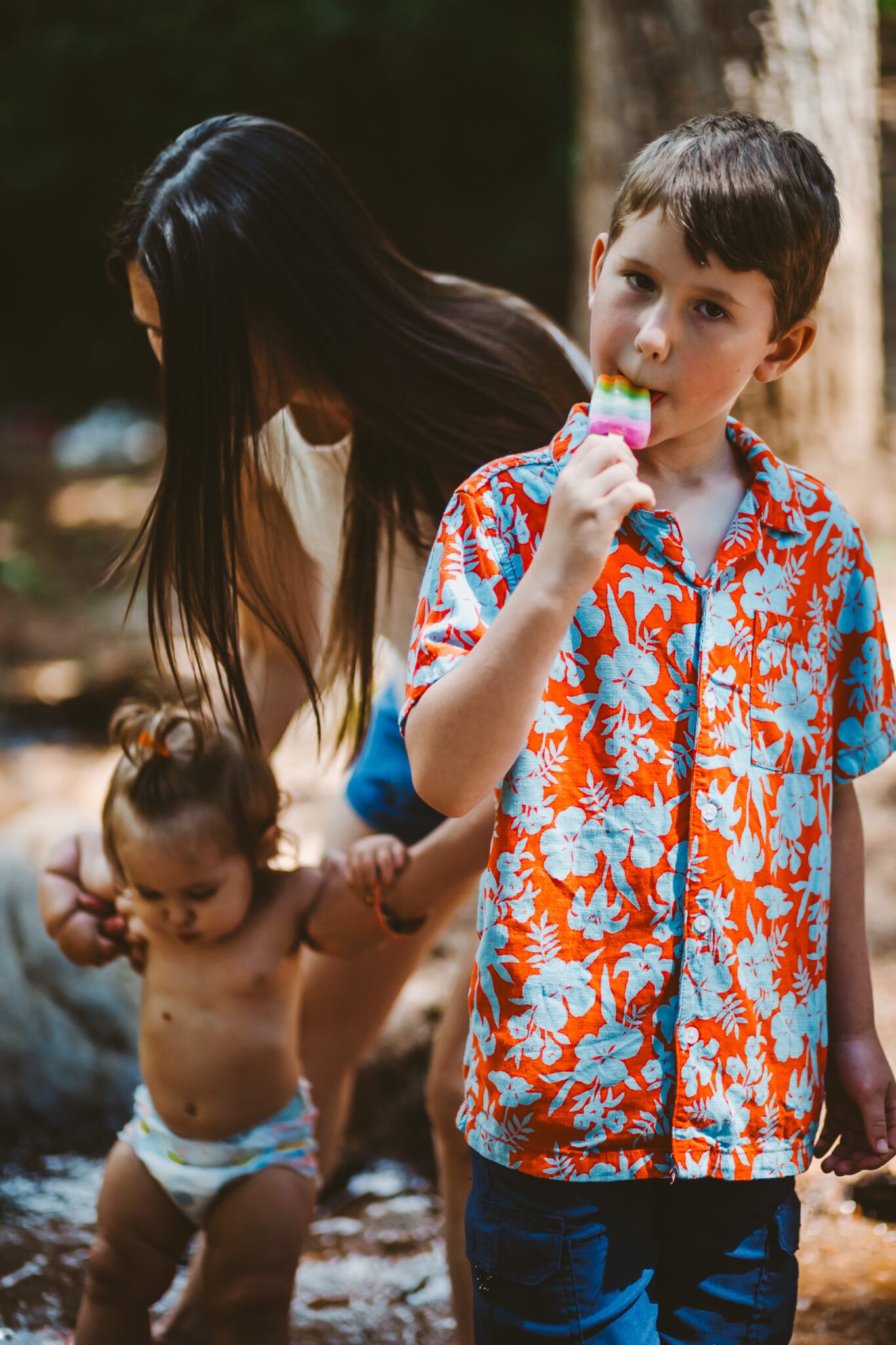 young boy eating popscicle with mother and toddler playing in creek behind