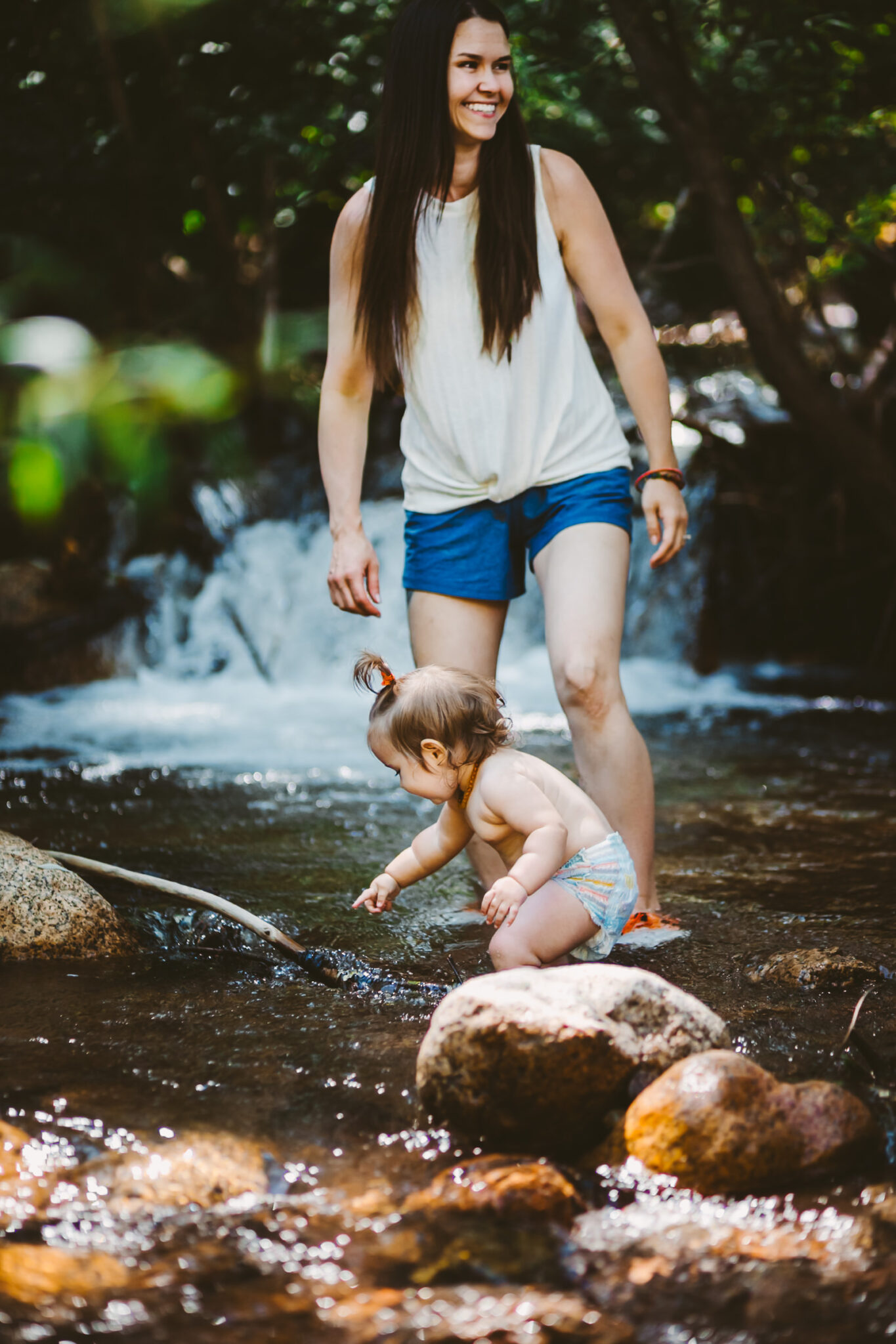 toddler plays in creek at cheyenne canyon, mother smiles as she watches with waterfall in background