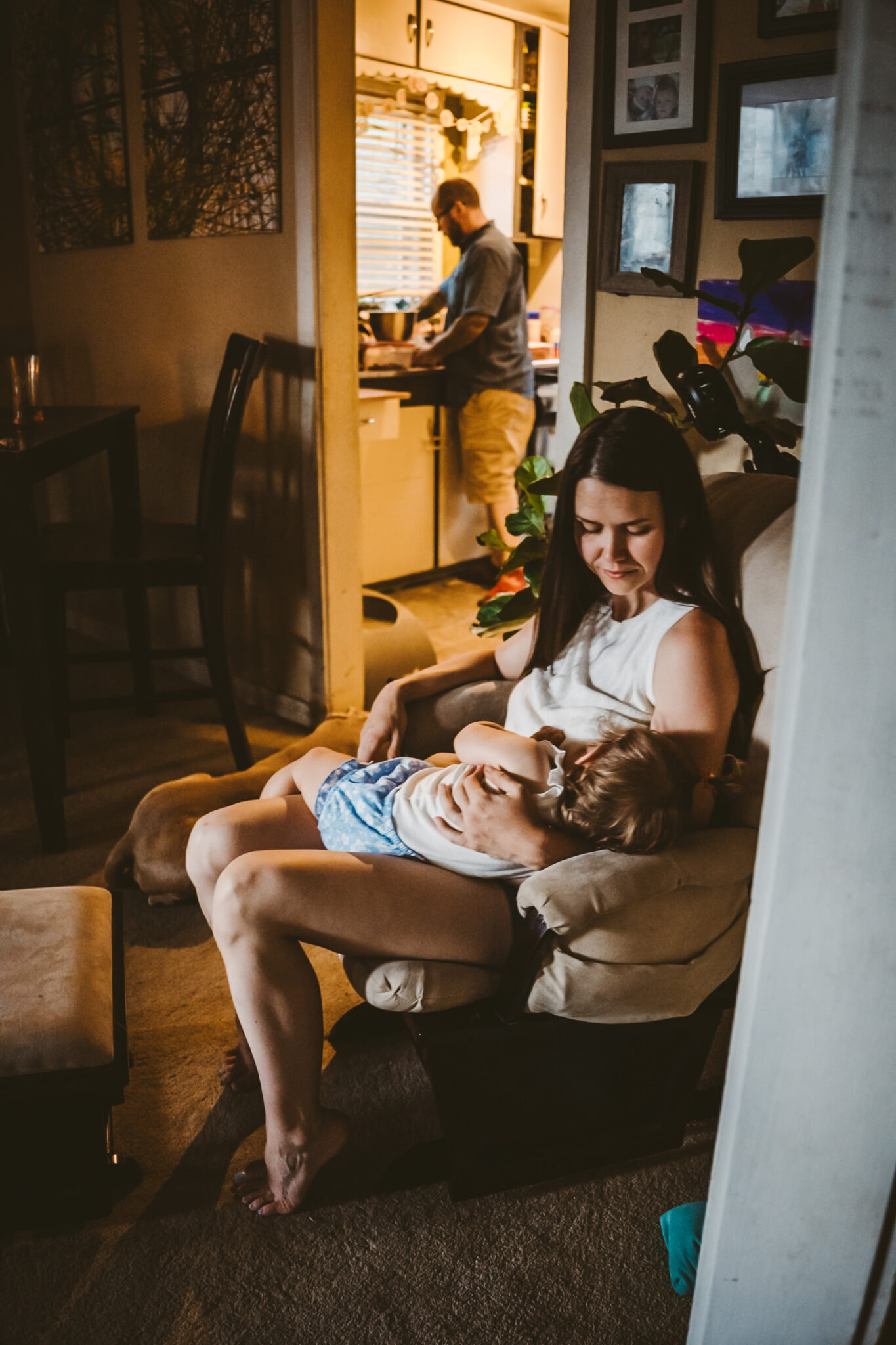 mother nurses toddler in living room while father makes dinner in the background